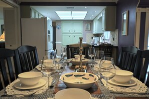 Een eetruimte met een tafel gedekt voor vier personen, met wit en blauw gemêleerde kommen, heldere wijnglazen en een decoratieve vaas in het midden.