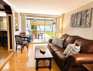 Een woonkamer met een bruine leren bank, een houten salontafel en een eethoek met stoelen en een tafel.