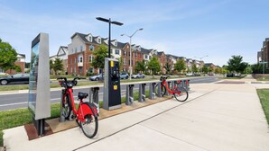 En cykeluthyrningsstation med en röd cykel och en laddstation i ett bostadsområde med hus och parkerade bilar.