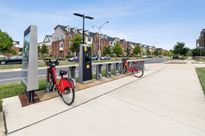 Eine Fahrradverleihstation mit einem roten Fahrrad und einer Ladestation in einer Wohngegend mit Häusern und geparkten Autos.