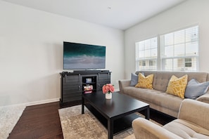A living room with a flat-screen TV mounted on a dark wooden cabinet, a beige sofa with patterned cushions, and a coffee table with a vase of flowers. - Your Oasis Away From Home, 4-bedroom townhome in Greenbelt (Greenbelt)