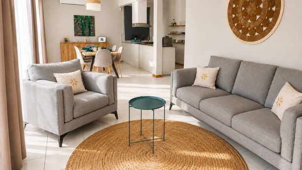 A modern living room with two grey sofas, a round woven rug, and a dining area in the background. - 3BR Sunset Sea View, Steps to Flic en Flac Beach (Flic en Flac)