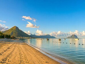 A beach with clear water, a mountain in the background, and boats. - 3BR Sunset Sea View, Steps to Flic en Flac Beach (Flic en Flac)