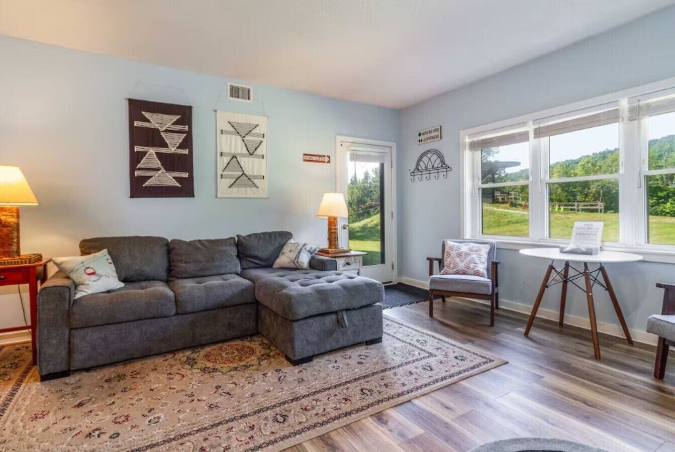 A living room with a sectional sofa, a round table, and a view of the outdoors through large windows.