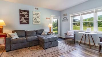 A living room with a sectional sofa, a round table, and a view of the outdoors through large windows.