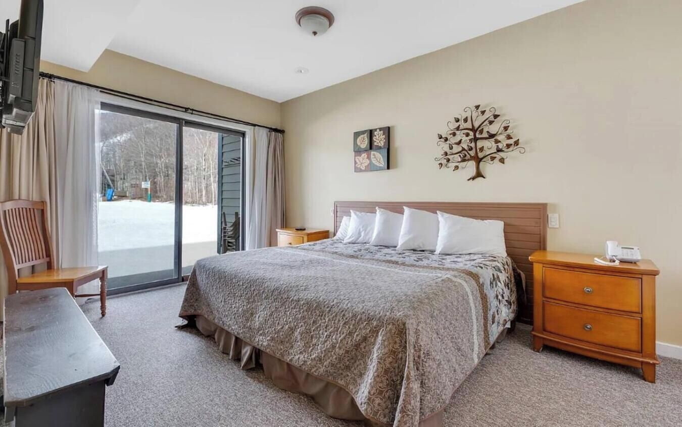 A hotel room with a large bed, a desk, a chair, a dresser, and a sliding glass door leading to a snowy outdoor area.