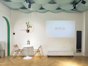 A modern room with a white wall, a round table with two chairs, a bench, a projector screen with 'NETFLIX' text, and ceiling lights. - Huge Balcony Home with Train Street View, Night Market, Max 6 (Hà Nội)