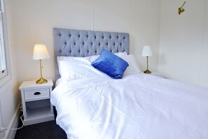 Una cama bien tendida con sábanas blancas y una única almohada azul, flanqueada por dos mesitas de noche con lámparas, en una habitación con un diseño sencillo y limpio.