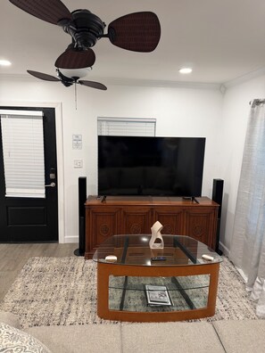A living room with a television, wooden entertainment unit, ceiling fan, and a glass coffee table. -  The Carlton — The perfect retreat to relax and unwind with a Great Location! (Tallahassee)