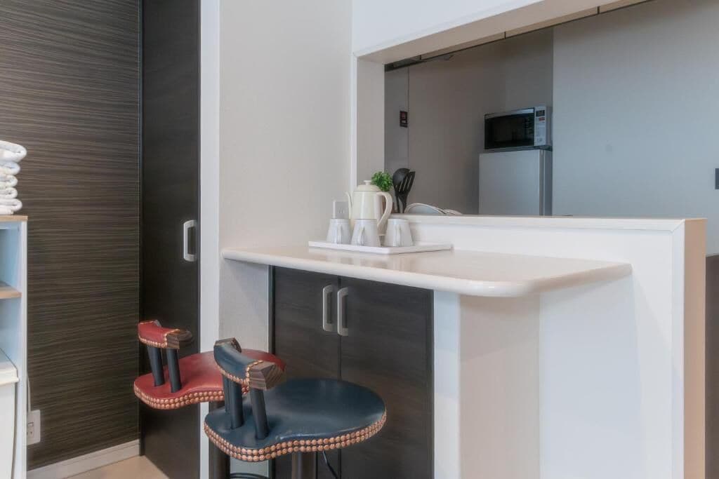 A compact kitchenette with a microwave, a small fridge, and a countertop with a kettle and cups. There are two stools with decorative elements.
