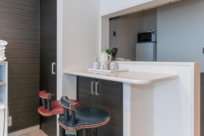 Une petite cuisine équipée comprenant un four à micro-ondes, un petit réfrigérateur et un plan de travail avec une bouilloire et des tasses. Deux tabourets décoratifs sont également présents.