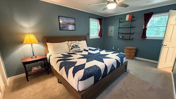 Ein Schlafzimmer mit einem Bett, einem Nachttisch, einer Lampe, einem Deckenventilator, einem Fenster mit Vorhängen und einem an der Wand befestigten Regal.