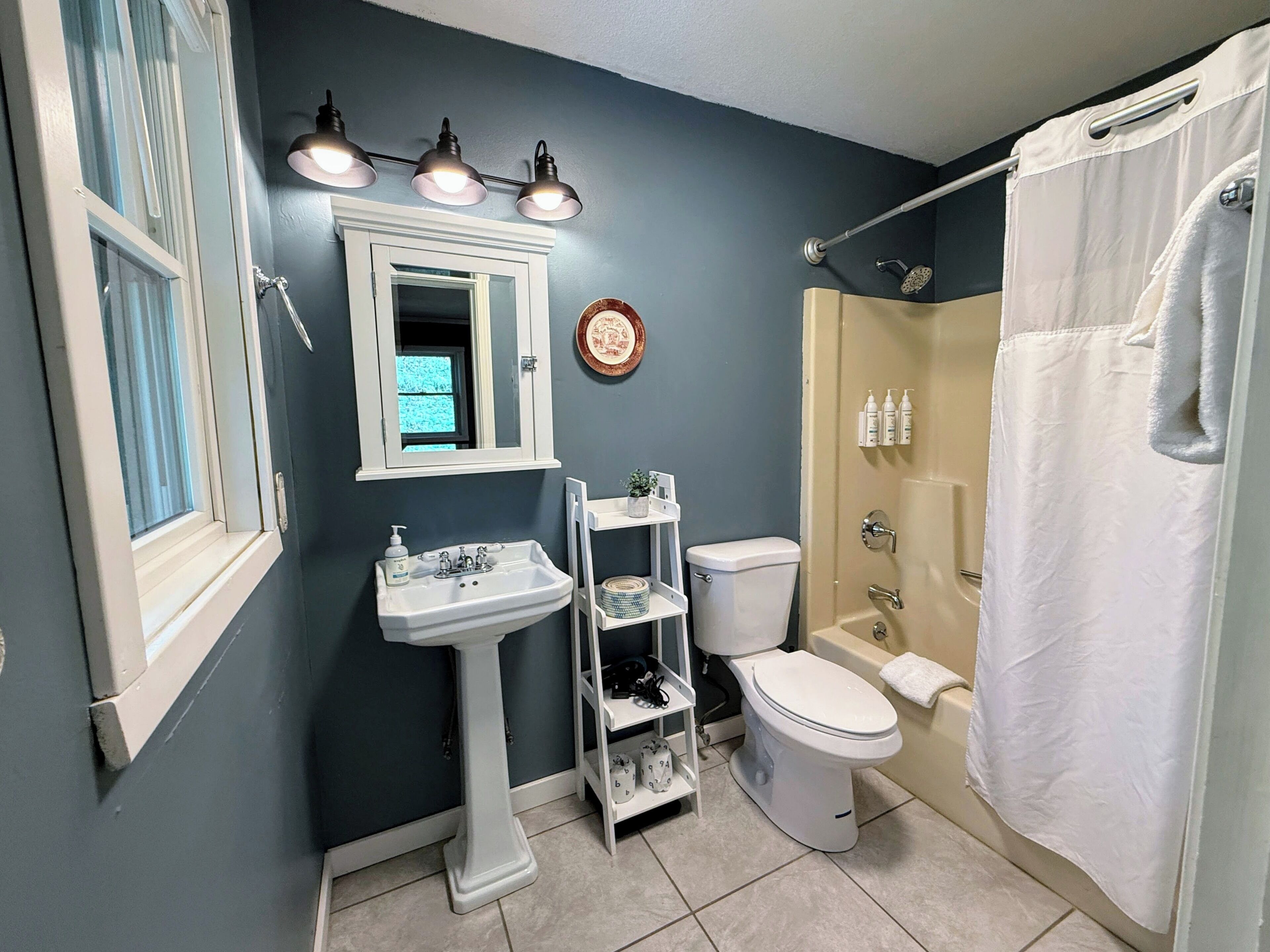 A bathroom with a pedestal sink, a toilet, a shower with a curtain, and a window with blinds.