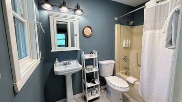 A bathroom with a pedestal sink, a toilet, a shower with a curtain, and a window with blinds.