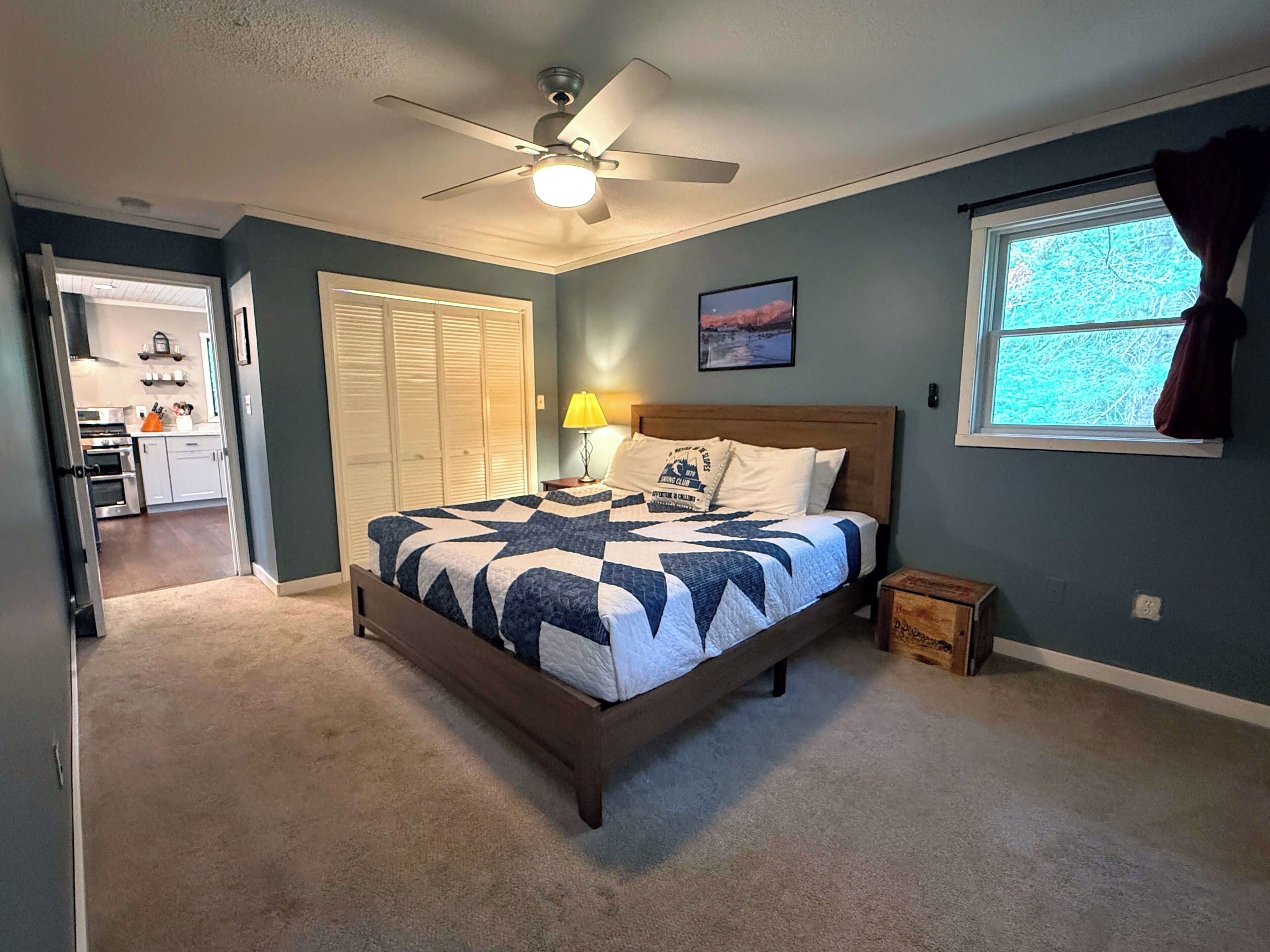 A bedroom with a bed, a ceiling fan, a window with curtains, a bedside lamp, and a door leading to a kitchen.