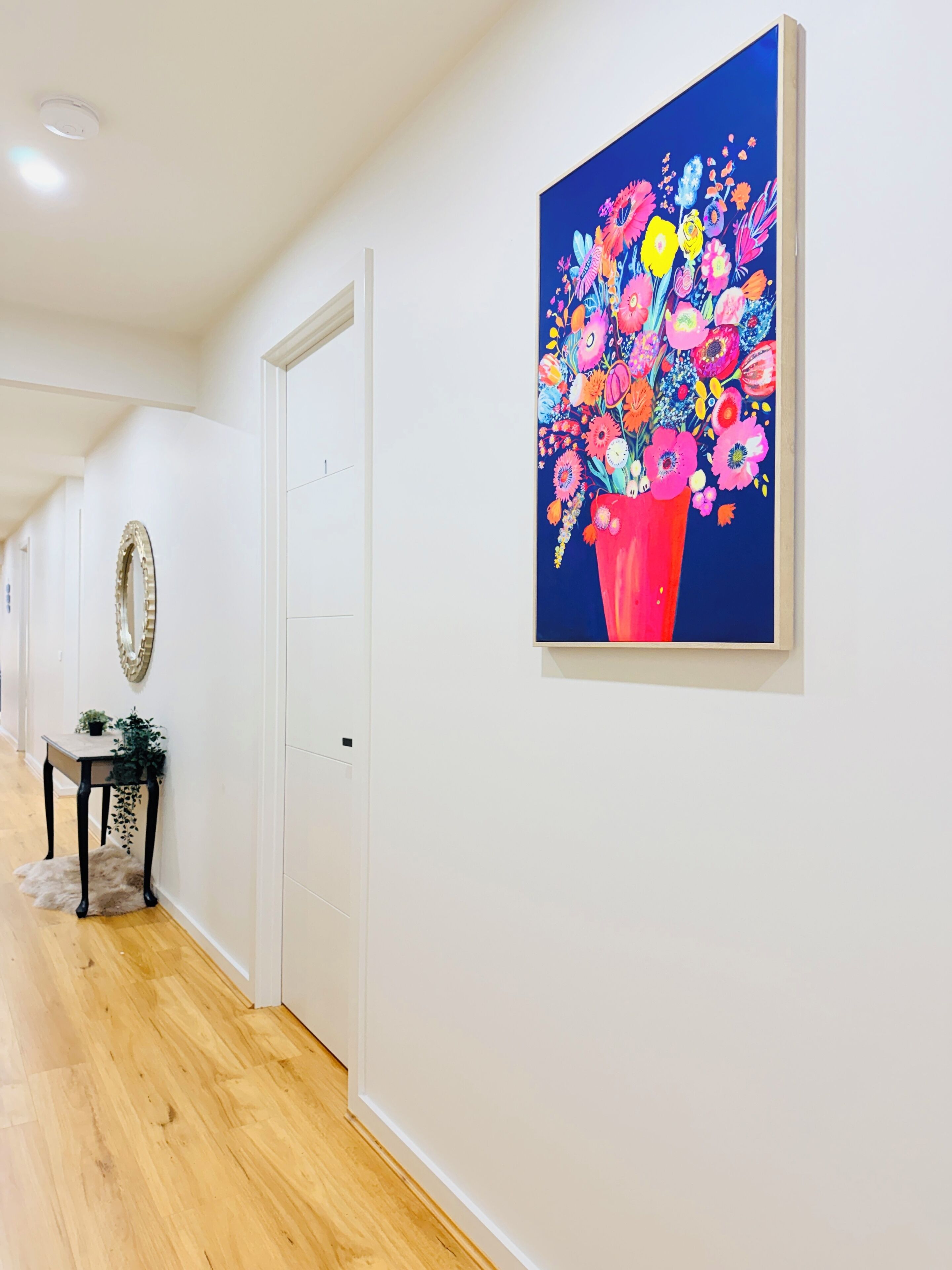 Un couloir avec une porte, un miroir, une petite table sur laquelle est posée une plante, et un tableau coloré accroché au mur.