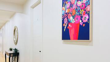 Un couloir avec une porte, un miroir, une petite table sur laquelle est posée une plante, et un tableau coloré accroché au mur.