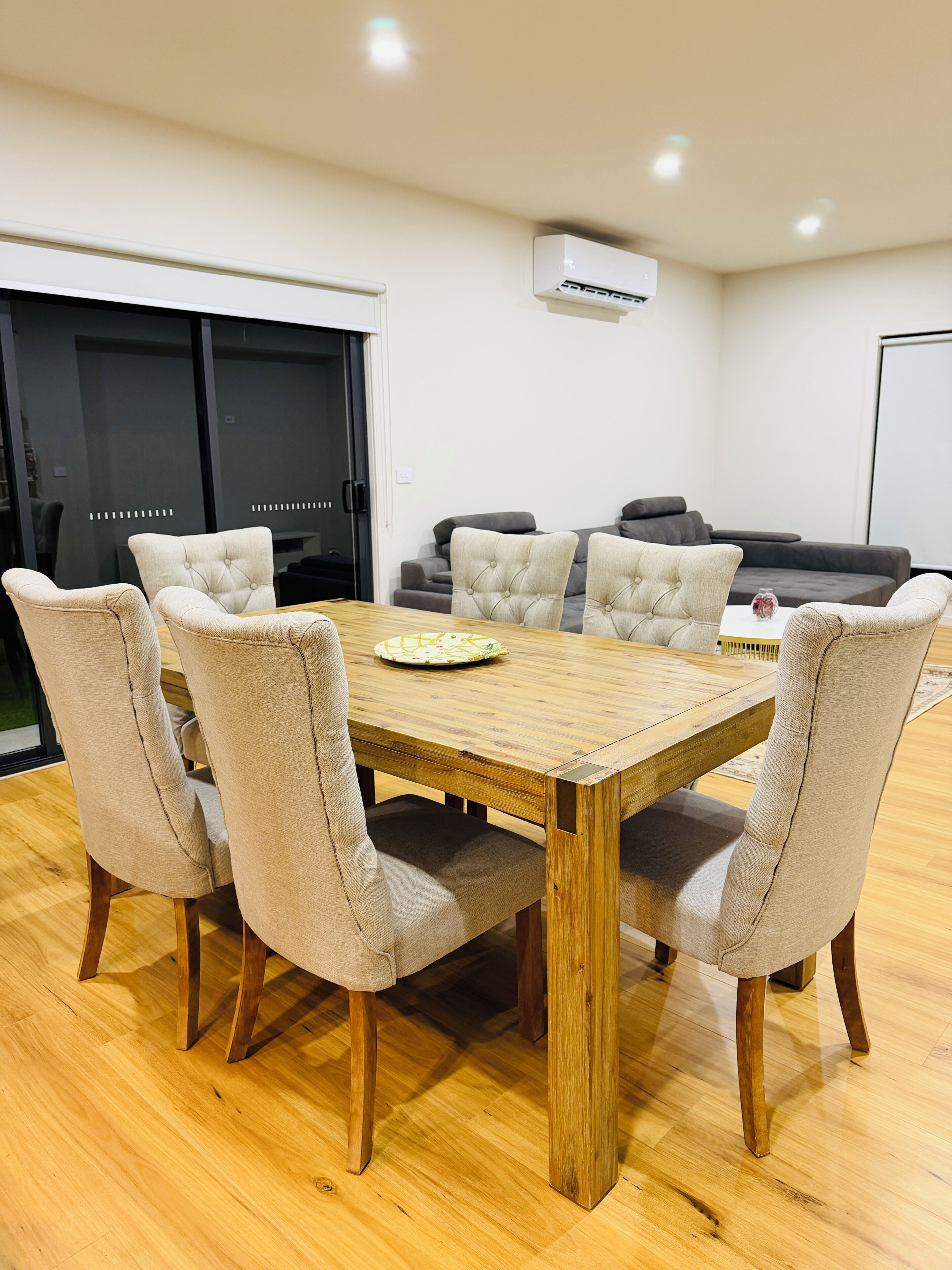 A dining area with a wooden table and six upholstered chairs, a black sofa, and a white air conditioning unit on the wall.