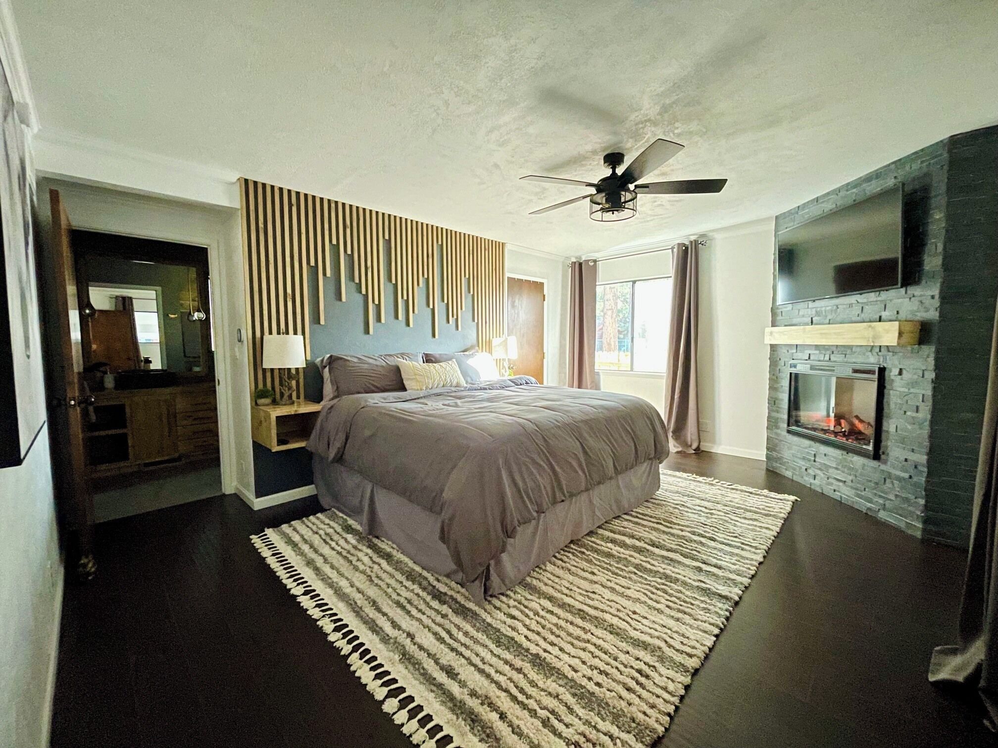 A modern bedroom with a bed, a ceiling fan, a fireplace, and a large window.