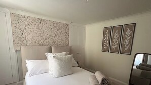 A neatly made bed with pillows and rolled towels, a headboard, and a wall with floral wallpaper and framed artwork. - 1 Bed - Sleeps 2 - Near Beach - Parking (Hastings)