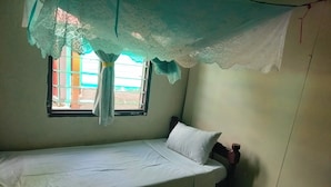 Un lit simple avec un matelas et un oreiller blancs, une fenêtre avec un rideau et une petite table de chevet sur laquelle se trouve une bouteille.