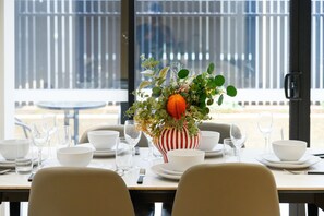 A dining table set with white plates, bowls, and glasses, with a striped vase containing greenery and an orange ornament.