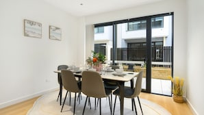 Un coin repas avec une table dressée pour quatre personnes, des chaises, un vase rempli de fleurs et une vue sur un bâtiment, visible à travers une porte vitrée.