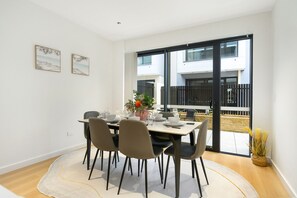 Un coin repas avec une table dressée pour quatre personnes, des chaises, un vase rempli de fleurs et une vue sur un bâtiment, visible à travers une porte vitrée.