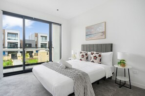 Une chambre à coucher moderne avec une grande fenêtre, un lit agrémenté d’oreillers à motifs floraux, une table de chevet avec une lampe et un tableau accroché au mur.