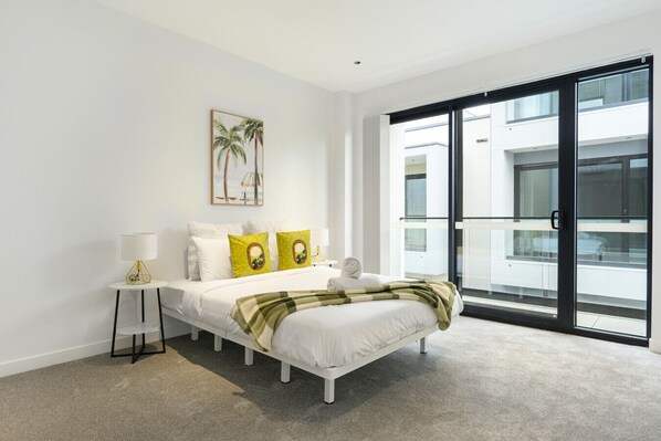 A modern bedroom with a bed, a bedside table with a lamp, a large window with a view of the building, and a painting on the wall.