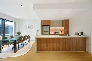 A modern kitchen with a central island, wooden cabinets, and a dining area with chairs and a table. - Brand New Family Home for 10 | Boxhill (Box Hill)
