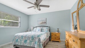 Ein Schlafzimmer mit Bett, Nachttisch, Kommode, Deckenventilator, Fenster mit Jalousien und einem Wandbild.