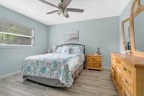 Ein Schlafzimmer mit Bett, Nachttisch, Kommode, Deckenventilator, Fenster mit Jalousien und einem Wandbild.