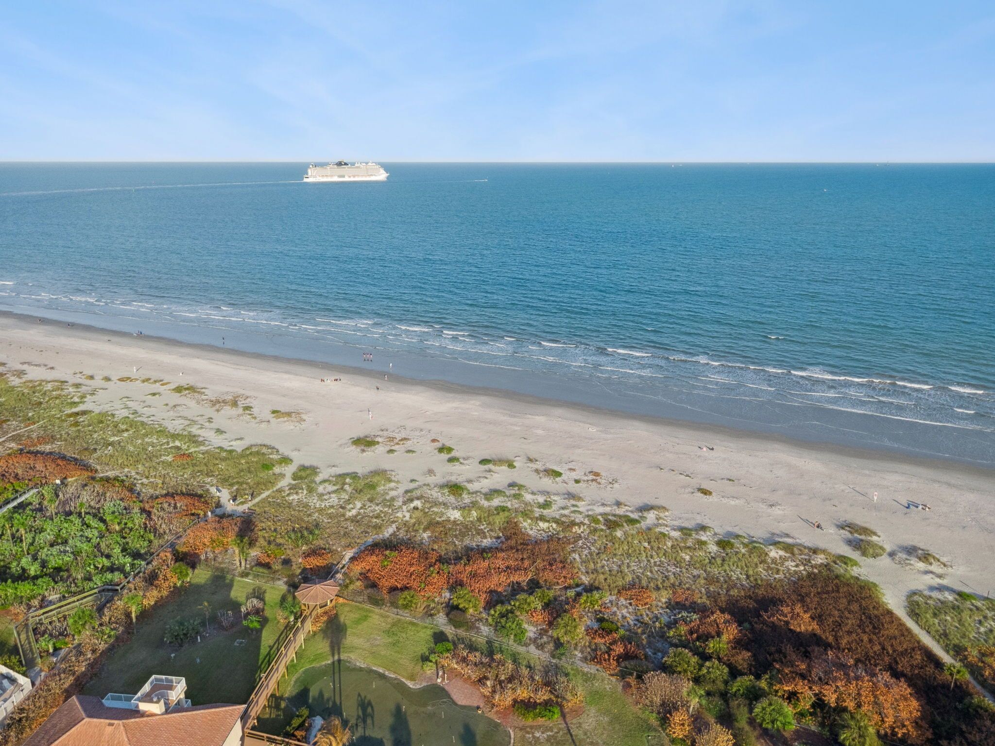 A coastal view with a cruise ship in the distance, sandy beach, and a residential area with vegetation.
