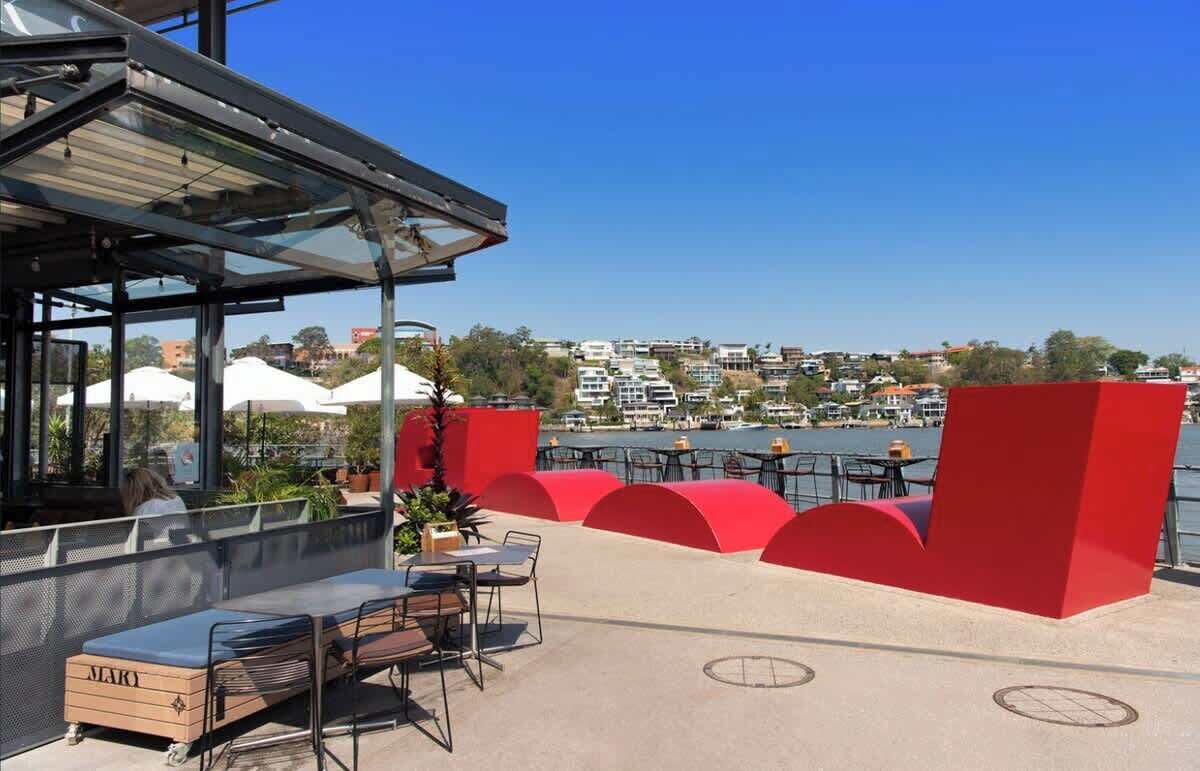 An outdoor seating area with tables and chairs, overlooking a body of water and a residential area.