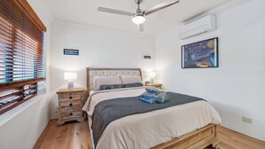 Un dormitorio con cama, mesitas de noche, ventilador de techo, unidad de aire acondicionado y una obra de arte en la pared.