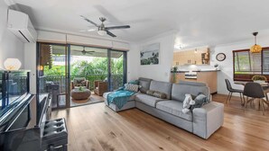 Una sala moderna con un sofá gris, un comedor con mesa y sillas, una cocina con armarios de madera y un balcón con mobiliario exterior.