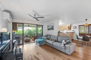 Ein modernes Wohnzimmer mit grauem Sofa, einem Essbereich mit Tisch und Stühlen, einer Küche mit Holdekor und einem Balkon mit Gartenmöbeln.