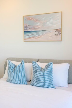 Ein Bett mit weißer Bettwäsche und zwei gemusterten Kissen, ein Strandbild an der Wand und ein grauer Kopfteil.