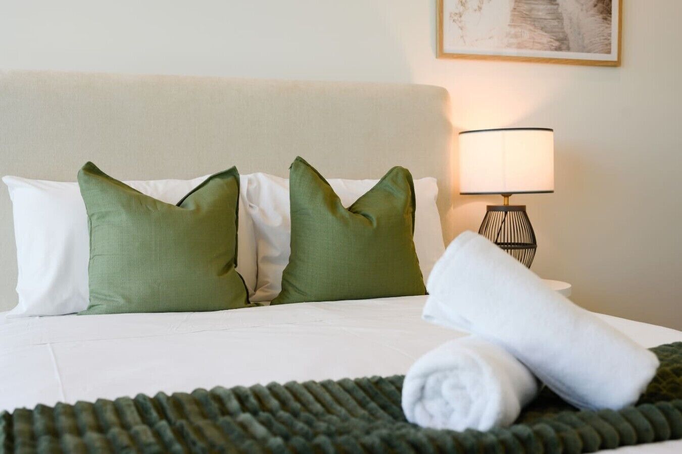 Una cama bien hecha con almohadas blancas y verdes, una toalla blanca enrollada y una manta verde.