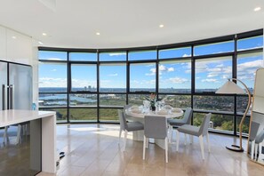 A modern dining area with a panoramic view of a cityscape and a river. - Spacious High-Rise 4Br Apartment | Olympic Park (Sydney Olympic Park)