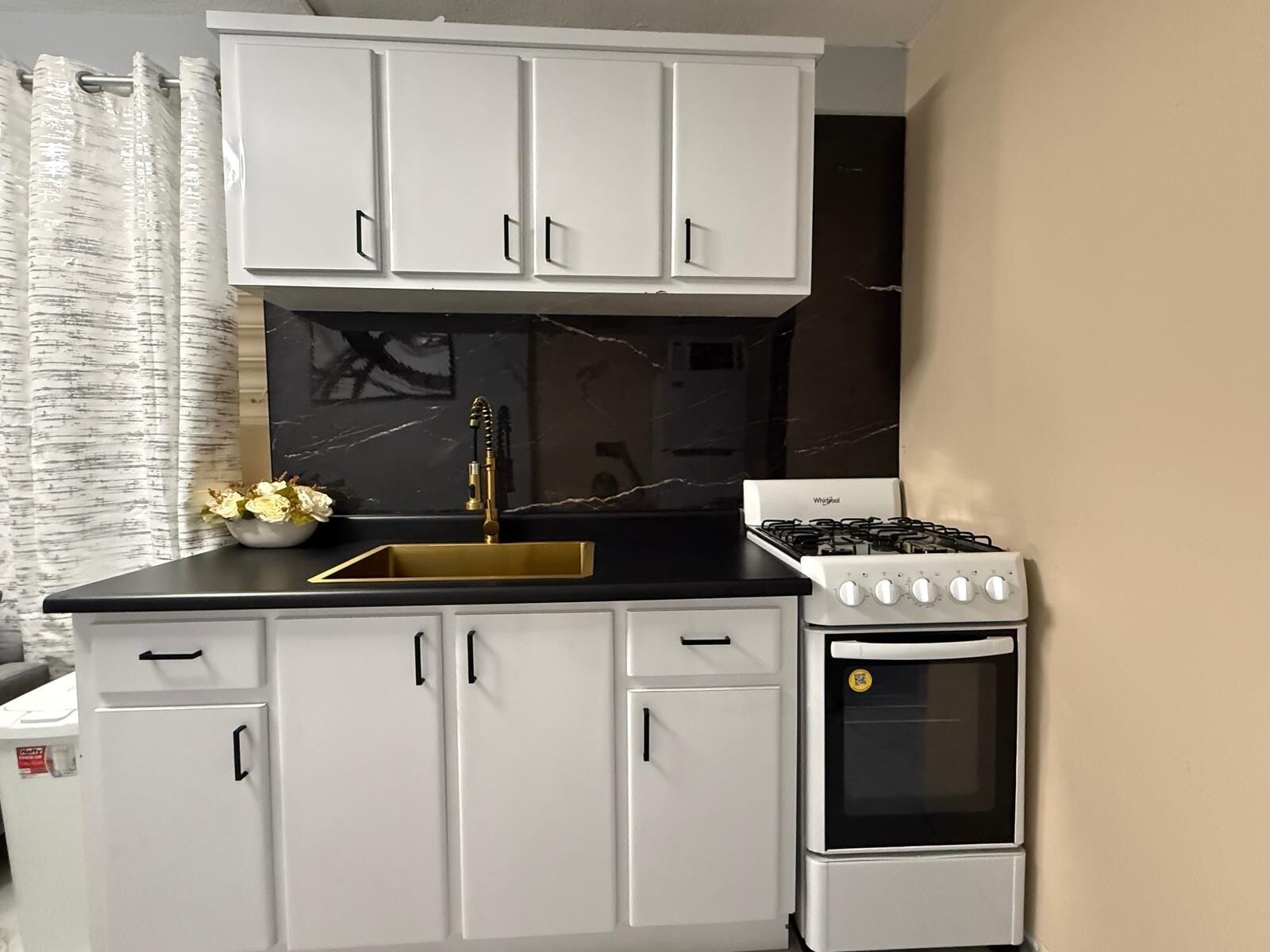Een kleine keuken met witte kastjes, een zwart aanrecht, een gouden kraan en een wit fornuis.
