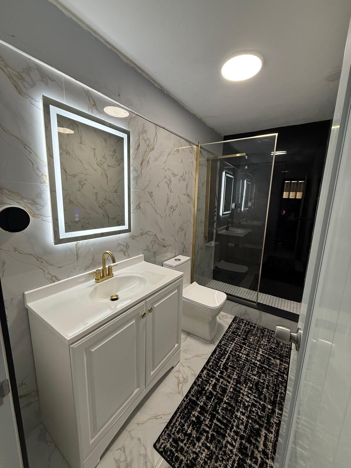 Ein modernes Badezimmer mit einem weißen Waschtisch, einem goldenen Wasserhahn, einem Spiegel, einer Toilette und einer Duschkabine aus Glas.