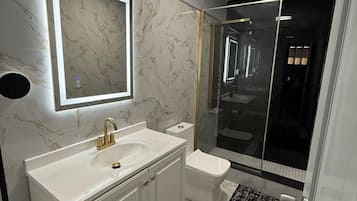 Ein modernes Badezimmer mit einem weißen Waschtisch, einem goldenen Wasserhahn, einem Spiegel, einer Toilette und einer Duschkabine aus Glas.