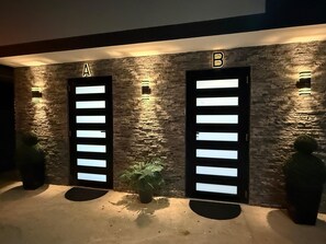 Dos puertas identificadas con las letras A y B, con paneles iluminados, sobre un muro de piedra texturizado, con macetas en la base.