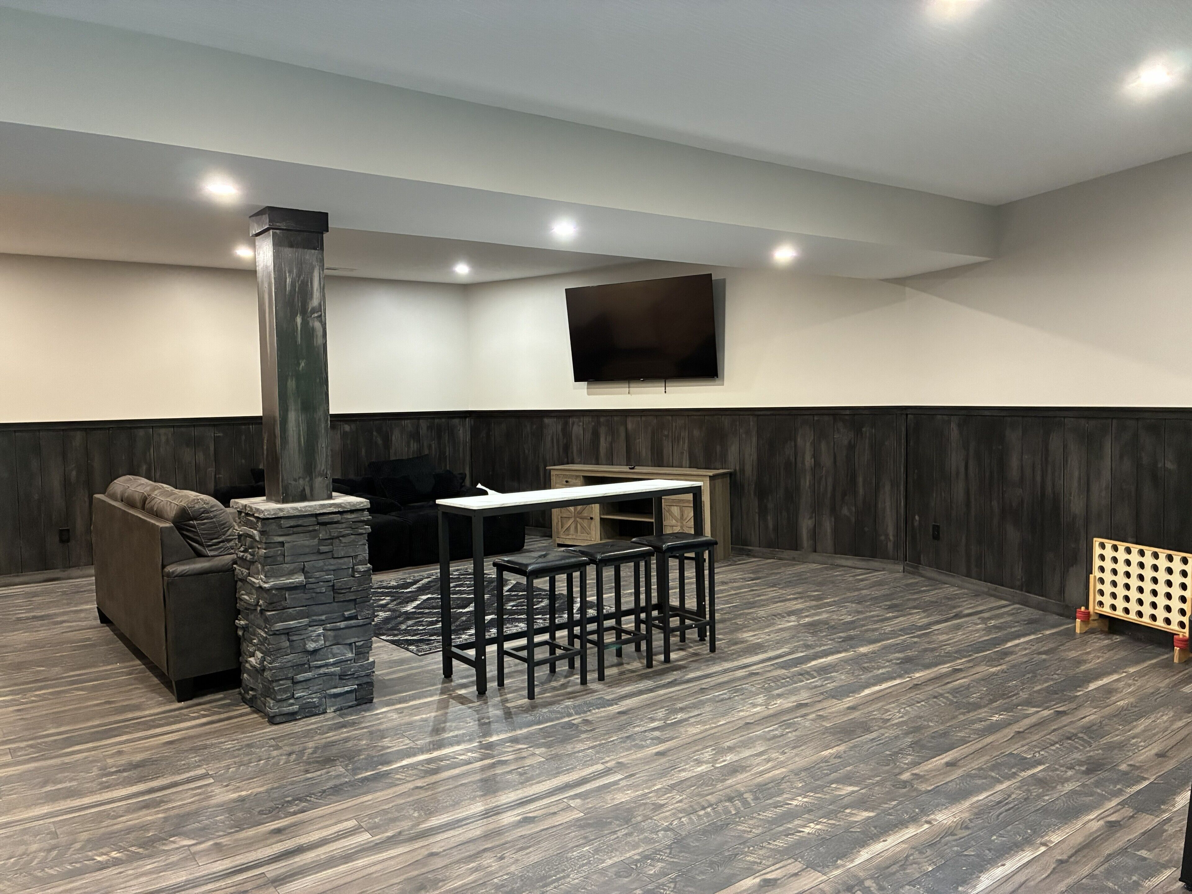 A modern living room with a stone fireplace, a black leather sofa, a dining table with stools, and a flat-screen TV mounted on the wall.