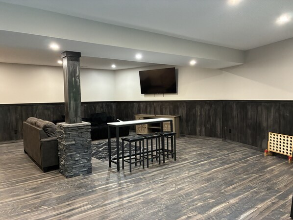 A modern living room with a stone fireplace, a black leather sofa, a dining table with stools, and a flat-screen TV mounted on the wall.
