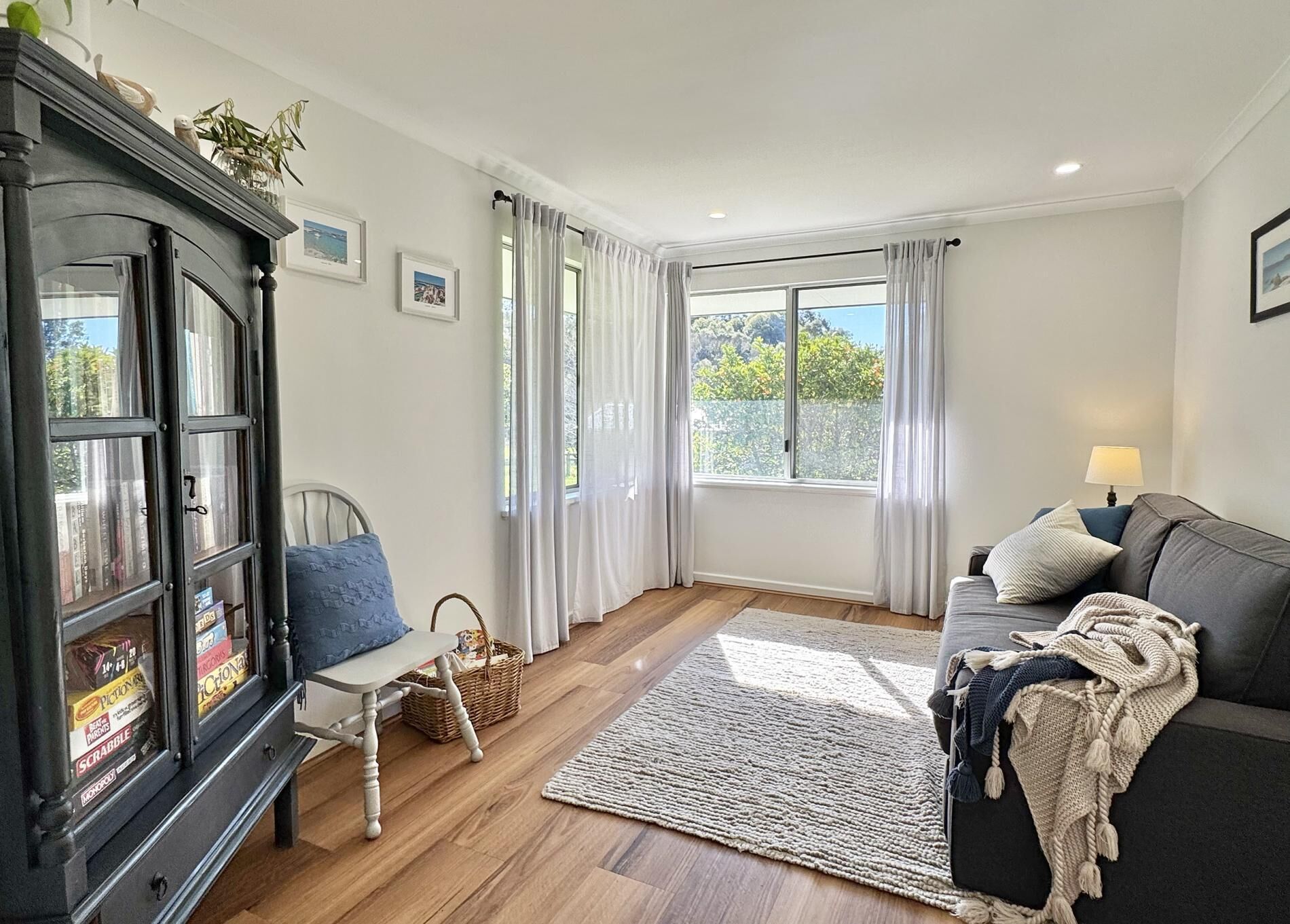 A cozy living room with a dark wooden cabinet, a white chair, a grey sofa, and a large window with curtains.