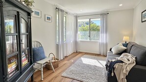 Ein gemütliches Wohnzimmer mit einem dunklen Holzschrank, einem weißen Sessel, einem grauen Sofa und einem großen Fenster mit Vorhängen.