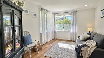 A cozy living room with a dark wooden cabinet, a white chair, a grey sofa, and a large window with curtains.